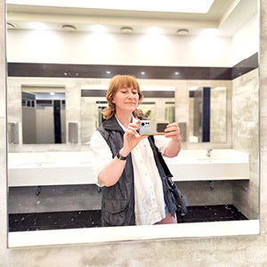 Frau macht im Bad ein Spiegelselfie mit dem Smartphone; hinter ihr sind Waschbecken, Spiegel und helle Wandfliesen zu sehen.
