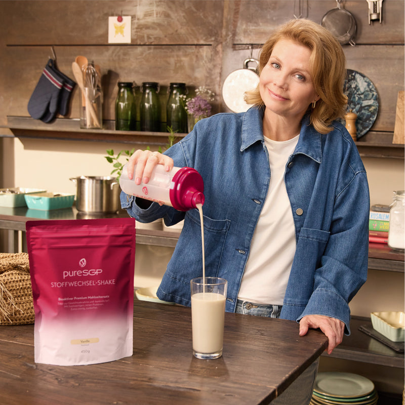 Annette Frier gießt in einer Küche einen cremigen Shake aus einem pinken Shaker in ein Glas; daneben steht ein Beutel pureSGP Stoffwechsel-Shake Vanille.