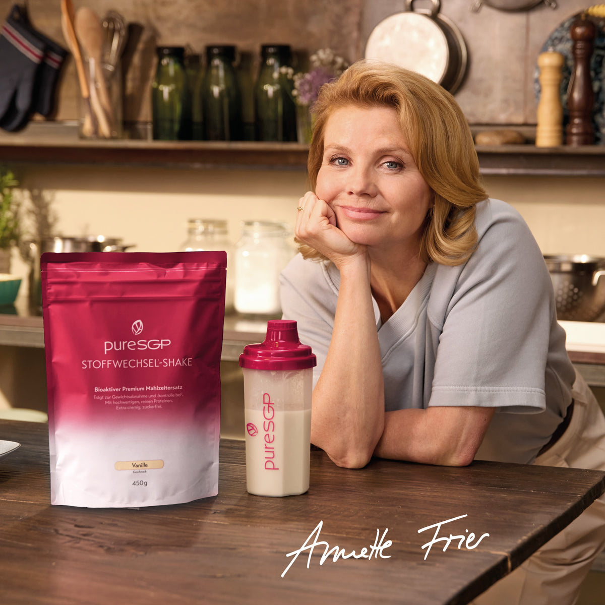 Annette Frier lehnt an einem Holztisch in einer Küche; daneben stehen ein Beutel pureSGP Stoffwechsel-Shake Vanille und ein gefüllter Shakebecher.